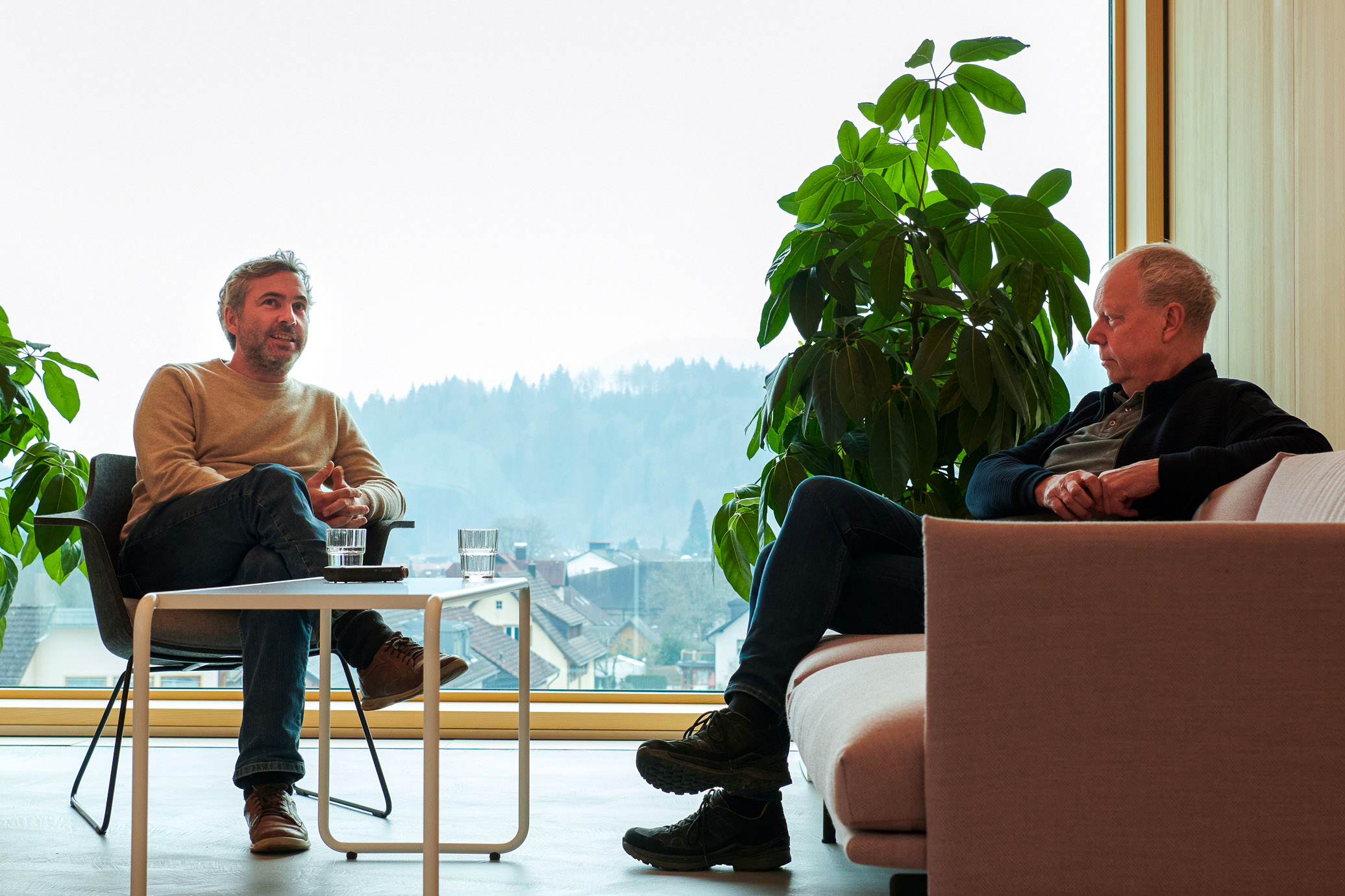 Vor einem großen Panoramafenster sitzen zwei Männer in entspannter Atmosphäre in einer Lounge-Ecke und sind ins Gespräch vertieft.