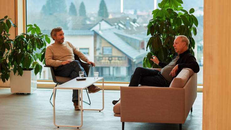 Vor einem großen Panoramafenster sitzen zwei Männer in entspannter Atmosphäre in einer Lounge-Ecke und sind ins Gespräch vertieft.