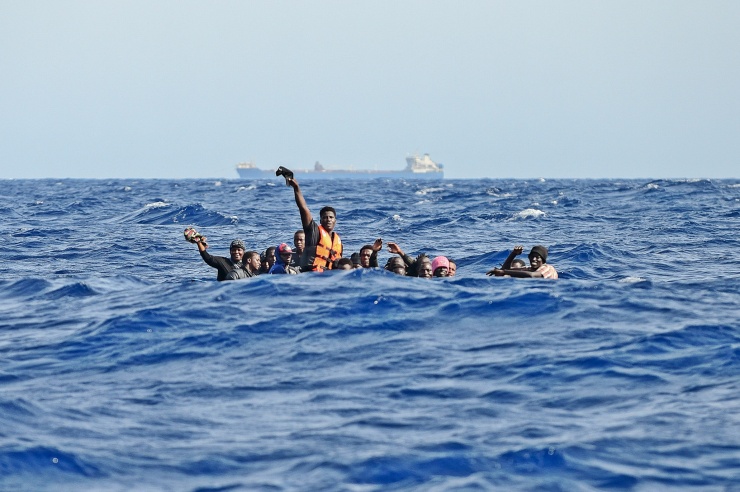 In den Wellen des aufgewühlten Meeres sind ein Dutzend winkende Flüchtende zu sehen, am Horizont sieht man ein großes Containerschiff.