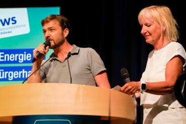 Ein Mann in grauem Polo-Shirt und eine blonde Frau in sommerlicher Kleidung stehen gemeinsam hinter einem Rednerpult.