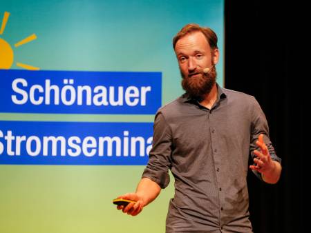 Vor der grünen Stele mt der Aufschrift "Schönauer Stromseminar" steht ein juner Mann mit roten Haaren und referiert.