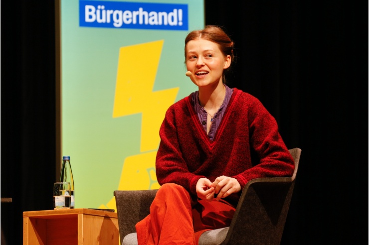 Eine junge Frau in weiter Kleidung und hochgesteckten Haaren spricht auf dem Podium.  