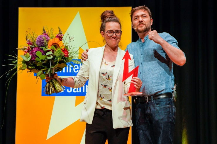 Eine Frau im hellen Blazer steht strahlend auf der Bühne, in der einen Hand den Award, in der anderen einen Blumenstrauß.