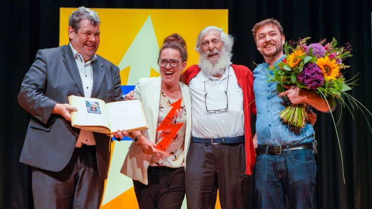 Vor der gelben Stele stehen Bürgermeister Schelshorn mit Katja Diehl, Michael und Sebastian. Die drei umarmen sich und alle lachen herzlich.