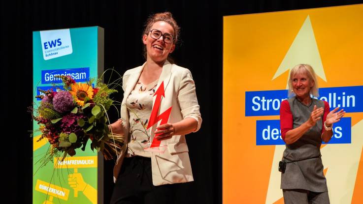 Eine Frau im hellen Blazer steht strahlend auf der Bühne, in der einen Hand den Award, in der anderen einen Blumenstrauß.