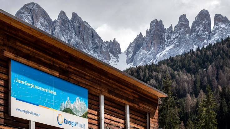 Hinter einem Holzhaus erstreckt sich ein Hang voller Tannen, aus dem eine zerklüftete Gipfelkette emporragt. 