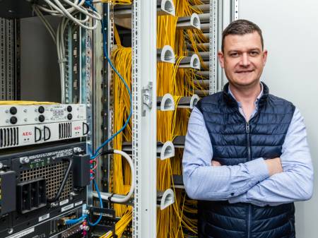  A middle-aged man wears a blue waistcoat while standing next to a switch box with his arms folded. 