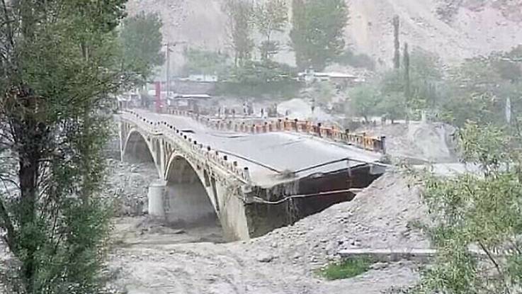 Eine Steinbrücke steht stark beschädigt über einem Flusslauf – der Straßenbelag ist gewellt, die Zufahrt gekappt.