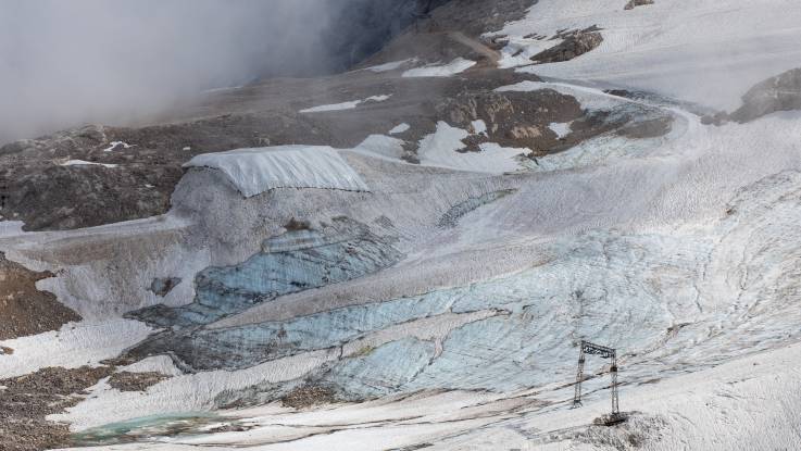 Der Hang ist großflächig schnee- und eisfrei, ein Schneehaufen ist mit weißer Plane abgedeckt. Im Vordergrund steht ein Schlepplifts. 