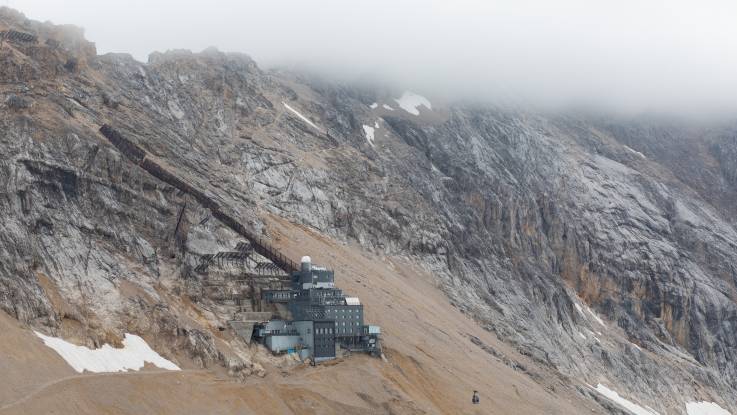 Am Hang einer massiven Gebirgswand steht ein einzelnes graues Bauwerk, zu dem eine Seilbahn führt. 