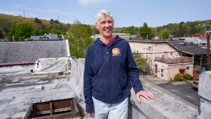 Eine Frau mit kurzen grauen Haaren, ausgewaschener Jeans und blauem Kapuzenpulli steht lächelnd auf einem Dach.