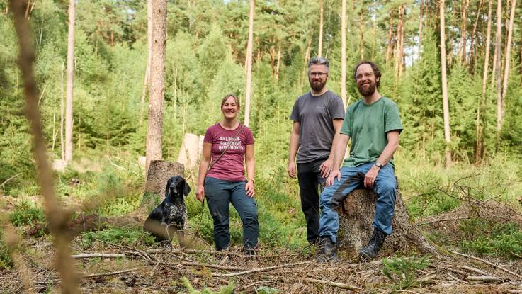 Zu der Dreiergruppe auf der Lichtung hat sich ein Hund gesellt – die Zweibeiner lächeln in die Kamera.