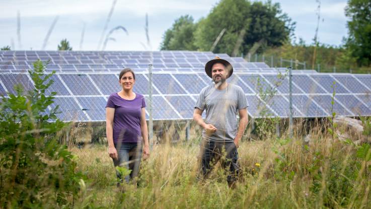 Ein Mann und eine Frau stehen lächelnd auf einer Wildwiese – im Hintergrund schieben sich PV-Module und Bäume ins Bild.