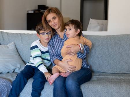 A woman sits next to a schoolchild on a couch while she has a toddler on her lap.