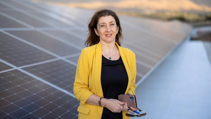 A woman in a yellow blazer stands smiling in front of a PV system, which is out of focus in the background.