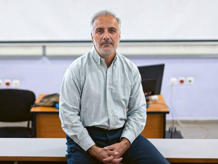 A grey-haired man sits on a table with his hands in his lap and a whiteboard behind him. 