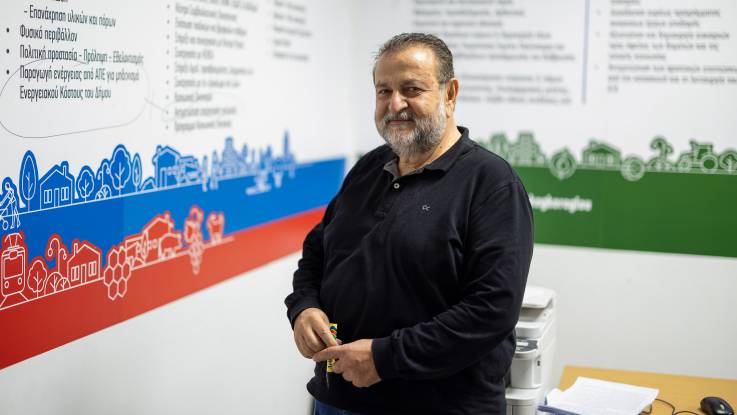 A man with a gray beard stands next to an information board and holds a thick marker in his hand. 