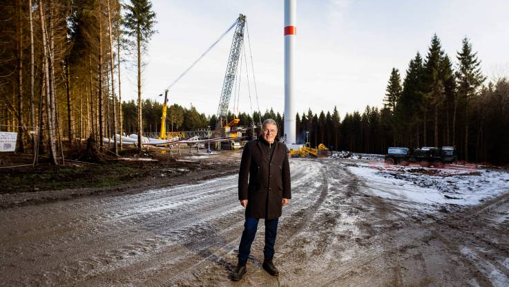 Der Mann mit Brille und Zopf steht vor einer Baustelle im Wald, hinter ihm wird ein Windrad mithilfe eines Krans errichtet. 