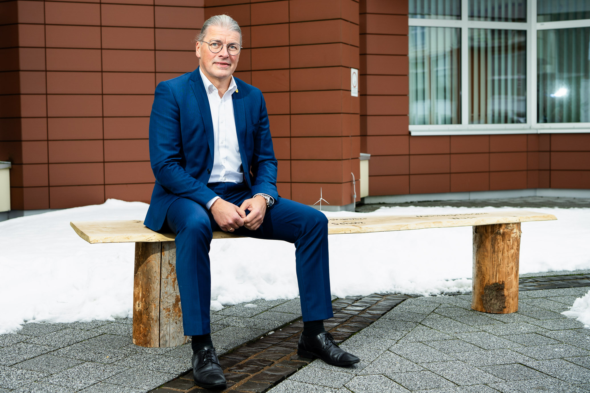 Ein Mann mit Brille und grauen, zum Zopf zusammengebundenen Haaren, sitzt im Anzug auf einer Bank.