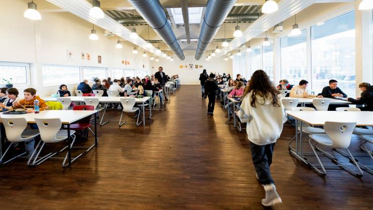 In einem hellen langen Raum mit dunklem Parkett sitzen zahlreiche Kinder zu Tisch und essen Mittag. 