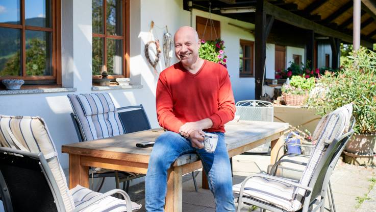 Der Mann mit dem roten Pullover Glatze lächelt noch immer, während er mit einer Kaffeetasse auf einem großen Tisch vor einem Haus sitzt.  