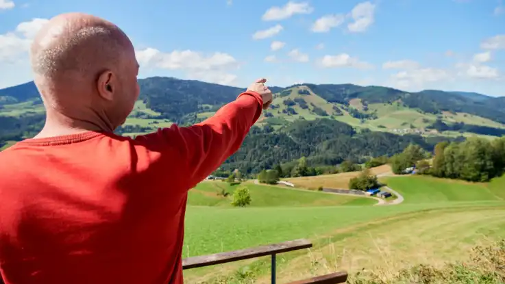 Ein Mann im roten Pullover und spärlich werdendem Haar ist von hinten zu sehen, der mit seinem Zeigefinger auf einen Bergkamm in der Ferne deutet. 