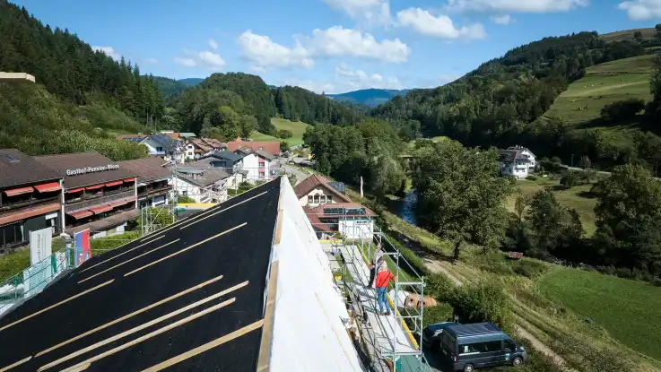 Ein angeschrägtes Dach ist noch nicht komplett eingedeckt – auf einem Gerüst daneben stehen zwei Personen.  