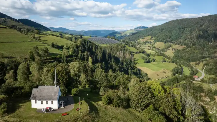 : Im Vordergrund steht eine kleine Kapelle mit weißen Mauern und Zierturm – deutlich dahinter lässt sich die PV-Anlage erkennen.