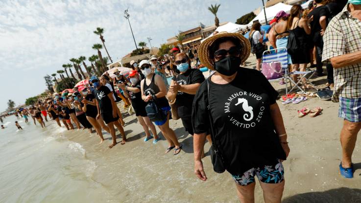 Entlang eines Strandes stehen unzählige Menschen mit Gesichtsmaske und Badebekleidung mit den Füßen im Meer.