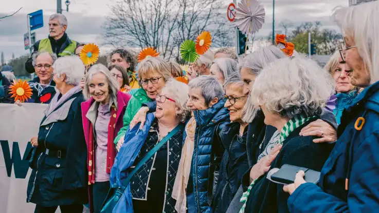 Eine Gruppe älterer Personen liegt sich freudig in den Armen – einige halten ein Transparent, andere Papierblumen. 