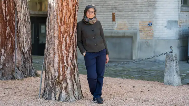 Die Frau mit der Kurzhaarfrisur steht nun im Freien an einem Baum gelehnt und schaut freundlich zur Kamera. 