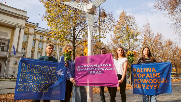 Eine Gruppe junger Menschen hält drei Banner mit verschiedenen Aufschriften, zwischen ihnen steht eine Windrad-Attrappe.