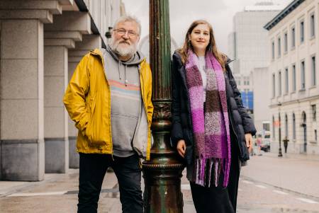  Ein Mann mit grauem Haar und eine jüngere Frau mit hellbraunem Haar lehnen sich an einer grünen Laterne an. 