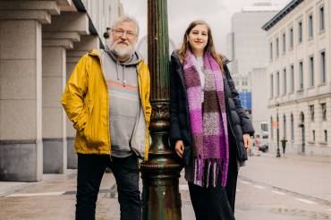 Ein Mann mit grauem Haar und eine jüngere Frau mit hellbraunem Haar lehnen sich an einer grünen Laterne an. 