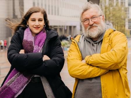 Eine junge Frau und ein älterer Mann stehen im Wind, verschränken demonstrativ freundlich die Arme und schauen in die Kamera.