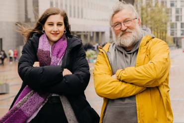 Eine junge Frau und ein älterer Mann stehen im Wind, verschränken demonstrativ freundlich die Arme und schauen in die Kamera.