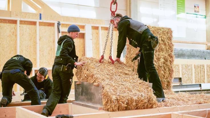 In der Werkhalle arbeiten vier Zimmerleute, zwei von ihnen pressen einen Strohballen in das am Boden liegende Wandelement aus Holz.