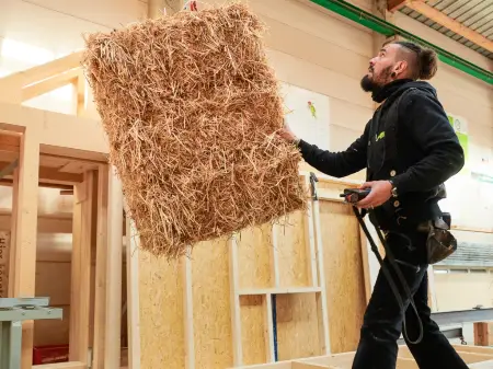 Ein Zimmermann manövriert einen Strohballen, der von der Decke herab an einem Kran hängt. 