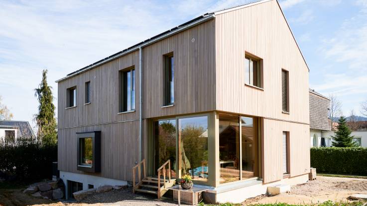 Ein fertiges Einfamilienhaus mit einer Holzverkleidung. Rundum sieht es noch sehr nach Baustelle aus.