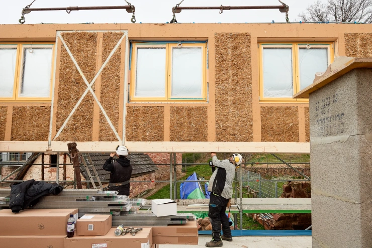 Eine vollständige Hauswand, inklusive eingebauter Fenster, schwebt über einem Hausfundament, und wird von zwei Bauleuten manövriert.