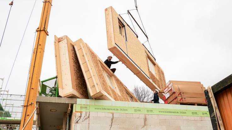 Auf der Baustelle hängt am Baukran ein Wandelement, das von einem Arbeiter neben bereits angelieferte Elemente platziert wird.