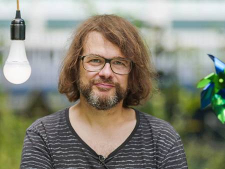 Ein Mann mit Brille, Bart und Zottelhaar hält eine Glühbirne in der Hand und schaut streng in Kamera.