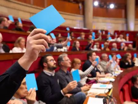 Im Halbrund eines Plenarsaals sitzen unterschiedliche Menschen und halten eine Abstimmung mit blauen Karten ab.