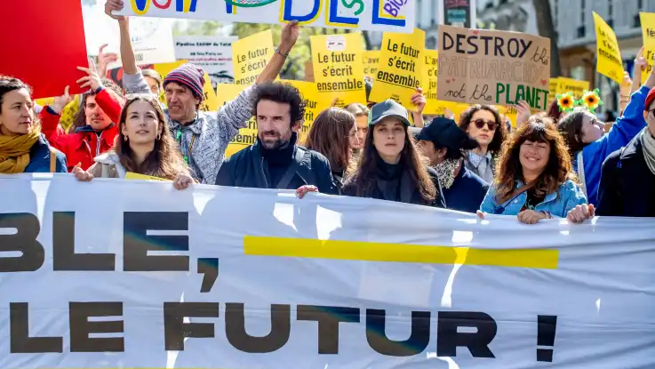 Eine Demo mit französischsprachigen Bannern, in der MItte hinter einem großen Transparent mit Aufschrift "...die Zukunft!" läuft ein dunkelhaariger Mann in den Vierzigern.