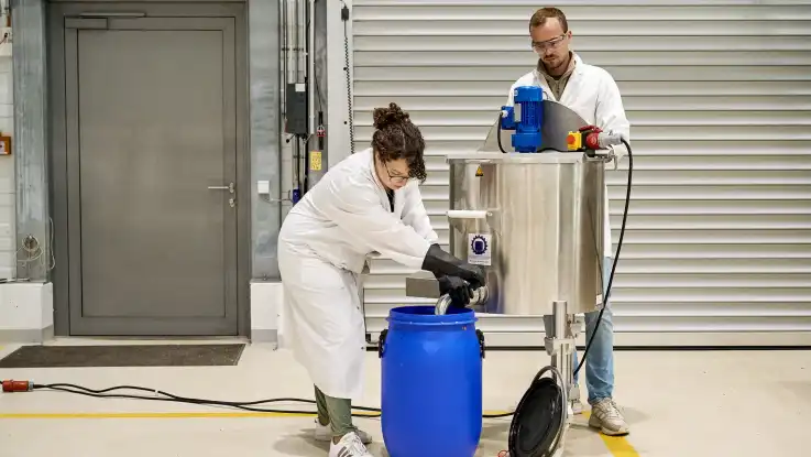 Zwei Personen in Laborkleidung stehen an einer zylinderförmigen Maschine, aus der ein breiter Hahn in ein blaues Kunststofffass mündet.