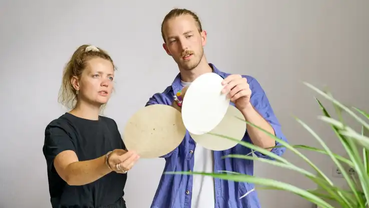 Zwei junge Erwachsene, ein Mann und eine Frau, betrachten drei kreisrunde Blätter aus Papier.