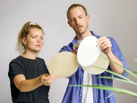 Zwei junge Erwachsene, ein Mann und eine Frau, betrachten drei kreisrunde Blätter aus Papier.