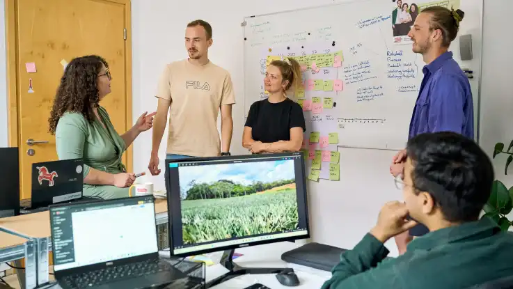 Besprechung im Büro: Während eine Person am Schreibtisch sitzt, stehen die anderen locker vor einem Whiteboard. 