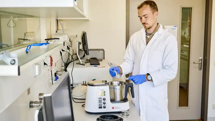 Ein junger Mann mit Kurzhaarfrisur trägt Kittel und blaue Schutzhandschuhe, während er im Labor arbeitet.