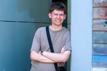 Ein junger Mann im T-Shirt steht mit verschränkten Armen angelehnt an eine Wand, die mit Graffiti besprüht.